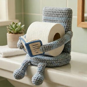 May include: A whimsical, crocheted toilet paper holder shaped like a seated figure, holding a roll of toilet paper and reading a small book. The holder is primarily blue and gray, with a white roll of paper. A small succulent plant sits nearby.