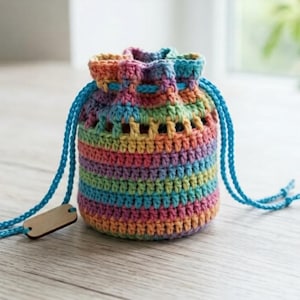 Puede incluir: Un bolso con cordón hecho a mano, tejido a ganchillo con los colores del arcoíris. El bolso tiene forma cilíndrica con rayas horizontales de varios colores. Tiene un cordón azul y una pequeña etiqueta de madera. El bolso mide unos 10 cm de alto.