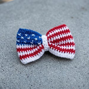 Pode incluir: Uma gravata borboleta de crochê no estilo da bandeira americana. A gravata borboleta apresenta uma seção azul com estrelas brancas e listras vermelhas e brancas, todas feitas com fio. Um acessório patriótico.