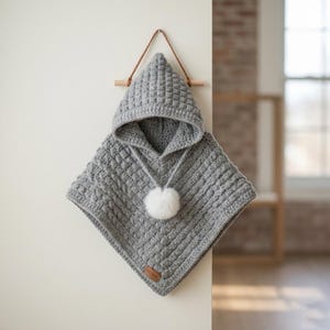May include: A gray, handmade crochet poncho with a hood and a white pom-pom. The poncho is hanging on a wooden bar with a leather strap. The textured knit design adds a cozy, handcrafted feel to this children's garment.