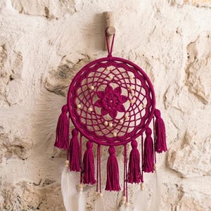 Pode incluir: Um apanhador de sonhos artesanal em um tom magenta profundo, apresentando um design de teia circular tecida com um motivo floral central. O apanhador de sonhos é adornado com borlas combinando e contas de madeira, e pendurado em um prendedor de madeira. Penas brancas pendem abaixo.