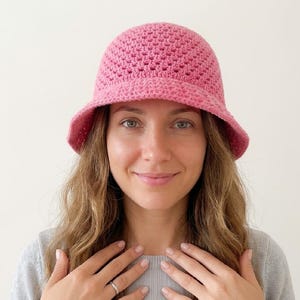 Puede incluir: Un sombrero de pescador rosa tejido a ganchillo con ala ancha. El sombrero está hecho de un hilo texturizado y tiene un tejido ligeramente abierto. El sombrero es de color rosa vibrante y liso. Es un accesorio elegante.