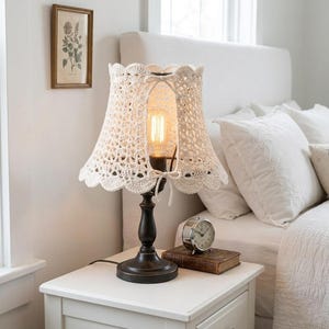 Puede incluir: Una pantalla de lámpara de ganchillo color crema con borde festoneado cubre una bombilla de estilo vintage. La lámpara está sobre una base marrón oscuro, colocada en una mesita de noche blanca. También hay un reloj pequeño y un libro.
