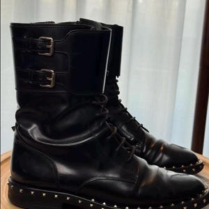 May include: A pair of black leather combat boots with silver-colored stud detailing around the soles. The boots have a lace-up design and two buckle straps at the top. The boots are displayed on a wooden surface.