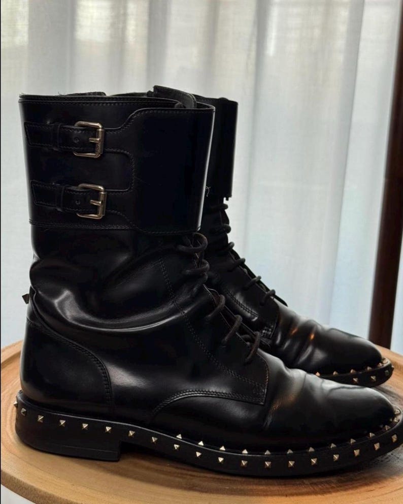 May include: A pair of black leather combat boots with silver-colored stud detailing around the soles. The boots have a lace-up design and two buckle straps at the top. The boots are displayed on a wooden surface.