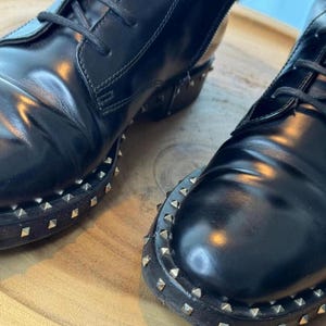 May include: Close-up of two black leather boots with laces. The boots have a glossy finish and are adorned with small, silver-colored studs around the soles. The boots are placed on a wooden surface.