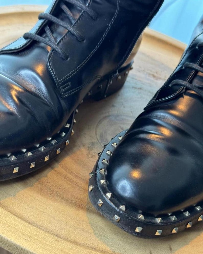 May include: Close-up of two black leather boots with laces. The boots have a glossy finish and are adorned with small, silver-colored studs around the soles. The boots are placed on a wooden surface.