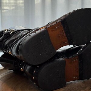 May include: Black leather boots with gold-colored stud accents. The boots have a buckle closure and a stacked heel. The soles are dark brown with a leather insert. The boots are on a wooden surface.