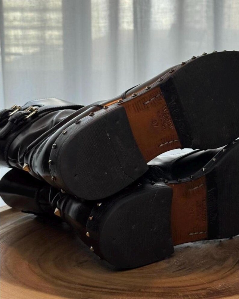 May include: Black leather boots with gold-colored stud accents. The boots have a buckle closure and a stacked heel. The soles are dark brown with a leather insert. The boots are on a wooden surface.