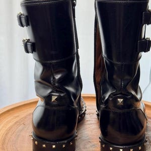 May include: Pair of black leather boots with gold-colored stud accents. The boots have two buckles on the upper shaft and a zipper on the side. The soles are also studded.