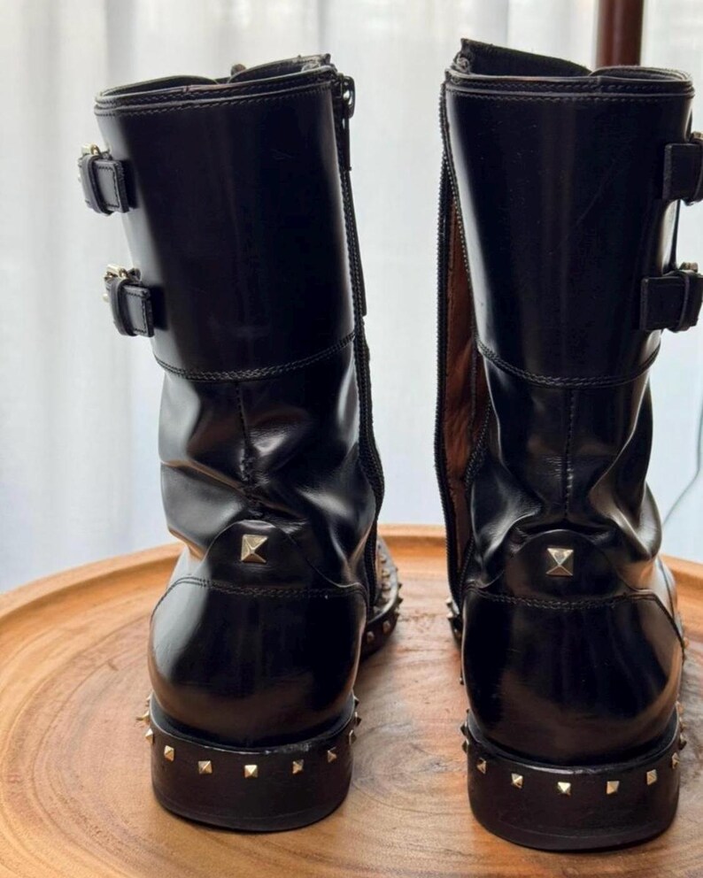 May include: Pair of black leather boots with gold-colored stud accents. The boots have two buckles on the upper shaft and a zipper on the side. The soles are also studded.