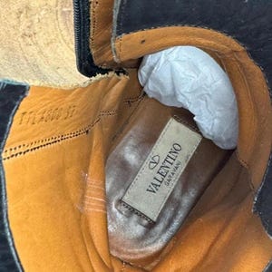 May include: Close-up of the interior of a black leather boot. The boot's interior is tan and features a label that reads "Valentino Garavani". The boot has a zipper and is stuffed with white paper.