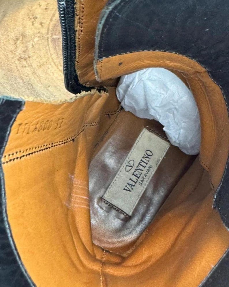 May include: Close-up of the interior of a black leather boot. The boot's interior is tan and features a label that reads "Valentino Garavani". The boot has a zipper and is stuffed with white paper.