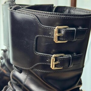 May include: Close-up of black leather boots with gold-colored buckles. The boots have a mid-calf height and feature two adjustable straps with buckles. The leather appears smooth and the stitching is visible.
