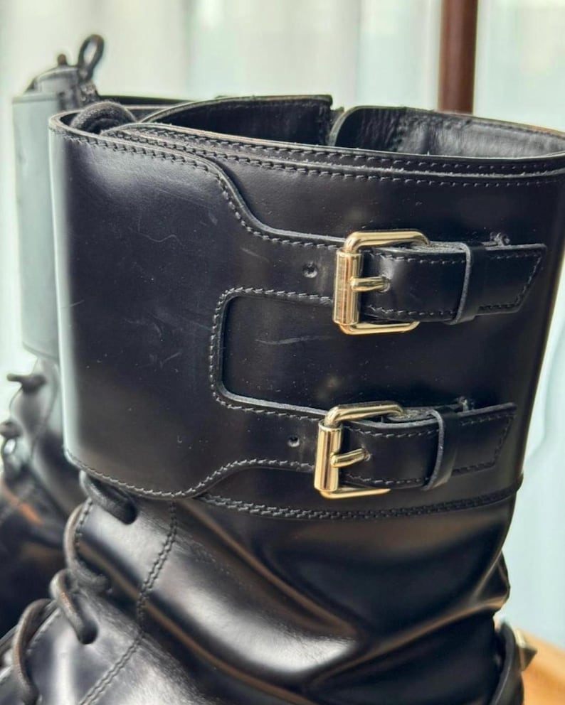 May include: Close-up of black leather boots with gold-colored buckles. The boots have a mid-calf height and feature two adjustable straps with buckles. The leather appears smooth and the stitching is visible.