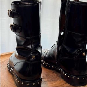 May include: Black leather combat boots with gold-tone stud detailing around the soles. The boots have a glossy finish, buckle accents, and a zipper on the side. The boots are displayed on a wooden surface.
