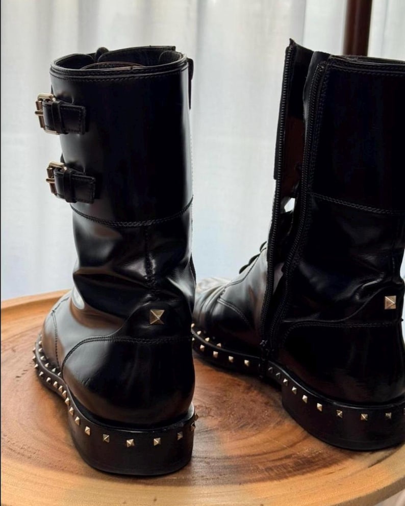 May include: Black leather combat boots with gold-tone stud detailing around the soles. The boots have a glossy finish, buckle accents, and a zipper on the side. The boots are displayed on a wooden surface.
