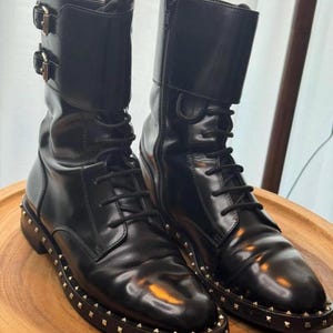 May include: A pair of black leather combat boots with a glossy finish. The boots feature a lace-up design, side zippers, and decorative buckles. The soles are adorned with small, gold-colored studs.