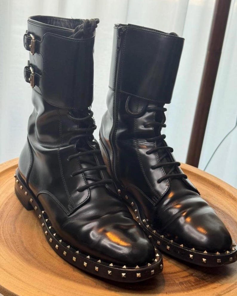 May include: A pair of black leather combat boots with a glossy finish. The boots feature a lace-up design, side zippers, and decorative buckles. The soles are adorned with small, gold-colored studs.