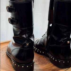 May include: Black leather boots with a glossy finish. The boots feature a studded sole and heel, along with decorative buckles and a side zipper. The boots are displayed on a wooden surface.