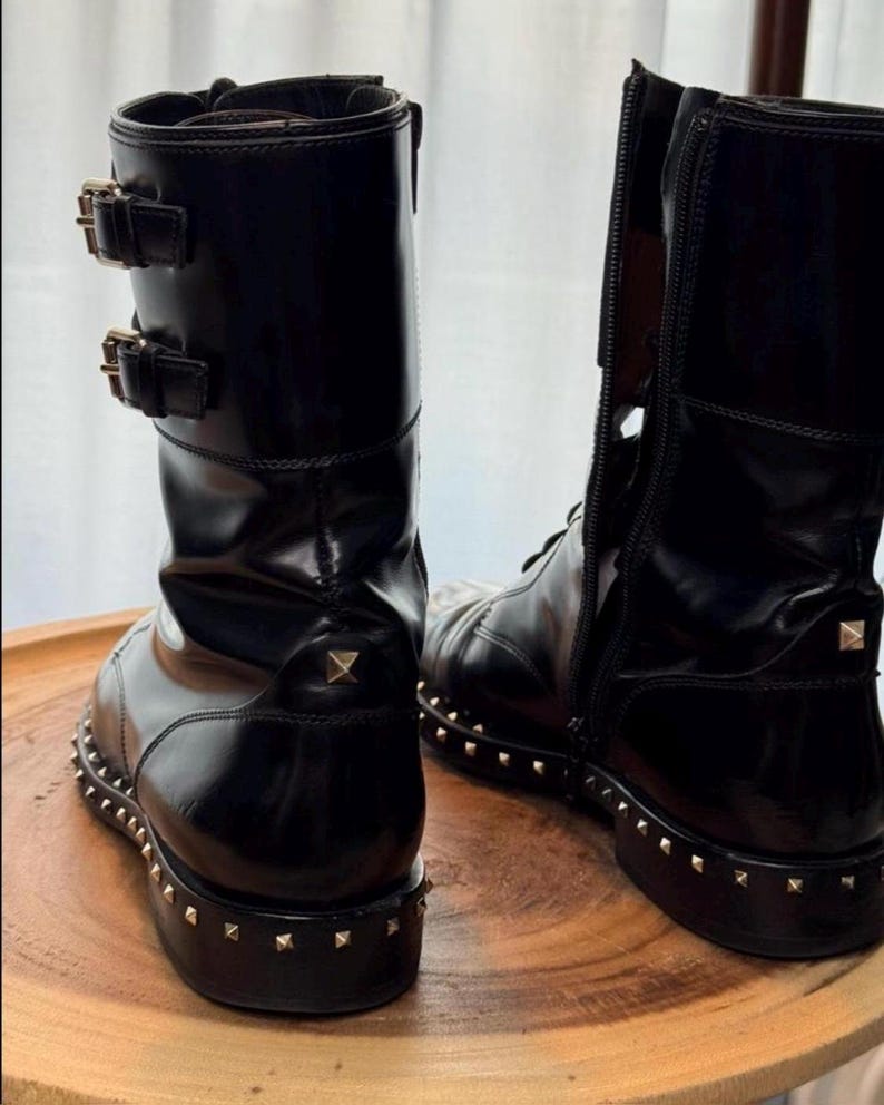 May include: Black leather boots with a glossy finish. The boots feature a studded sole and heel, along with decorative buckles and a side zipper. The boots are displayed on a wooden surface.