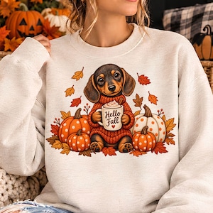 Puede incluir: Una sudadera beige de cuello redondo con un gráfico de un perro salchicha marrón con un suéter rojo, sosteniendo una taza blanca con el texto Hello Fall, rodeado de calabazas naranjas y blancas con lunares y hojas de otoño.
