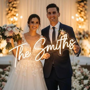 May include: A smiling couple poses at a wedding, holding a neon sign that reads "The Smiths." The bride wears a white wedding dress and carries a bouquet. The groom wears a navy suit. The background features floral arrangements and soft lighting.
