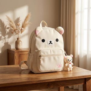 May include: A cream-colored backpack featuring a bear face design with rounded ears, black eyes, and a white muzzle. The front pocket displays a small pink paw print graphic. A matching miniature bear plush keychain hangs from the side zipper.