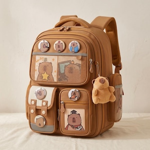 Pode incluir: Uma mochila castanha com múltiplos compartimentos com fecho, um bolso frontal de rede e vários pins circulares com desenhos de capivaras. Um porta-chaves de peluche de capivara pende do lado e os bolsos frontais exibem ilustrações de capivaras.
