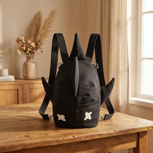 May include: A black backpack featuring a central dorsal fin, two side fins, and two white cross-shaped accents on the lower front. The bag includes adjustable black shoulder straps attached with silver-toned metal hardware and a structured main body.