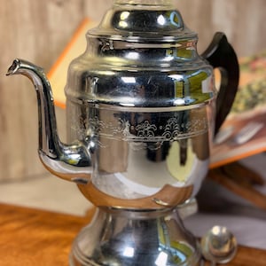 May include: A polished chrome coffee percolator featuring a curved spout, a black handle, and a clear glass knob on the lid. The body of the percolator is decorated with an etched horizontal band of scrollwork patterns around the center section.