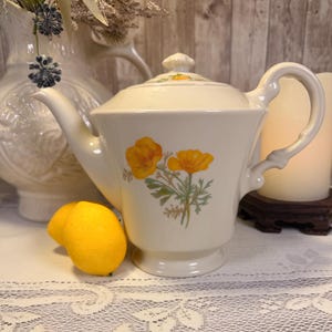May include: A cream-colored ceramic teapot featuring a floral pattern of two yellow poppies with green stems and leaves. The teapot has a matching lid with a small knob, a curved handle, and a tapered spout, positioned next to a bright yellow lemon.
