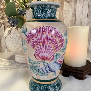 May include: A ceramic vase featuring a cream-coloured body with a hand-painted pink and purple seashell motif, green foliage, and dark green mottled patterns at the flared rim and base, accented by thin horizontal bands in a muted orange tone.