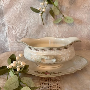 May include: A cream-colored gravy boat candle with a floral design, resting on a matching saucer. The candle has a single wick and is surrounded by decorative greenery. The set is placed on a neutral-toned surface.