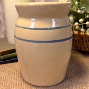 May include: A cream-colored ceramic crock featuring two horizontal blue stripes encircling the upper portion of the body. The vessel has a smooth, glossy finish, a wide opening, and a slightly tapered cylindrical shape with a rounded base.
