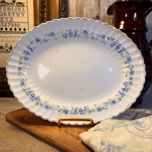 May include: An oval white ceramic platter with a scalloped, fluted edge. The rim features a continuous floral pattern of small blue flowers and green leaves. The center of the platter has a smooth, off-white surface with faint brown spots.