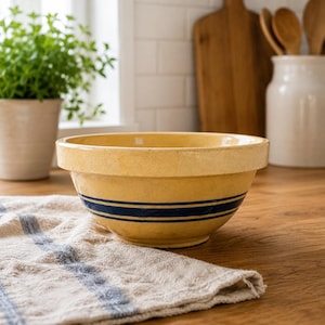 May include: A yellow ceramic mixing bowl featuring a wide, thick rim and two horizontal dark blue stripes encircling the exterior. The bowl has a smooth, glazed finish and a rounded base, presenting a classic design for kitchen preparation tasks.