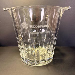 May include: A clear glass ice bucket featuring a textured, vertical ribbed pattern and two side handles. The front surface is etched with the Tupperware logo and the text Millennium Quest 2000. The vessel has a wide, open top and a sturdy base.
