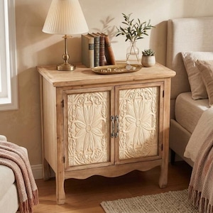 Pode incluir: Uma mesa de cabeceira de madeira clara com duas portas com um design floral em relevo. A mesa de cabeceira tem uma lâmpada, livros e itens decorativos em cima. As portas têm puxadores de metal.