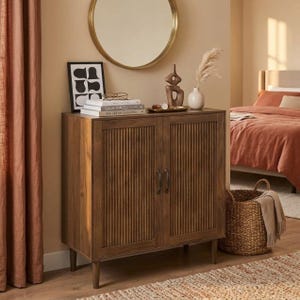 Puede incluir: Un mueble de madera con dos puertas y detalles de ranuras verticales. El mueble tiene un acabado marrón cálido y se apoya en cuatro patas cónicas. Un espejo redondo dorado cuelga encima del mueble y una cesta de mimbre está en el suelo.