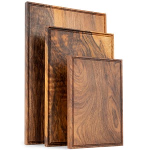 May include: A set of three rectangular wooden cutting boards of varying sizes, each featuring a distinct dark brown wood grain pattern and a recessed juice groove running along the perimeter of the top surface of every board.