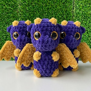 May include: Three plush, crocheted dragon toys. Each dragon has a deep purple body, large, round, black eyes, and small, yellow horns. The wings and feet are a soft, golden yellow. The dragons are standing on a white surface with a green background.