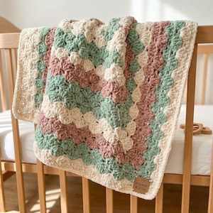 May include: A handmade crochet blanket in a granny square pattern. The blanket features alternating rows of cream, dusty rose, and sage green. The blanket is draped over a wooden cot, with a small tag that reads "HANDMADE WITH LOVE."
