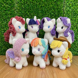 May include: A collection of seven handmade crochet ponies in various colors. Each pony has a white body, a colorful mane and tail, and blue eyes. Some ponies have wings or hats. The ponies are arranged in a group against a green background.