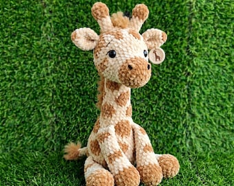 May include: A plush giraffe toy with a cream and brown spotted pattern. The stuffed animal has a long neck, small ears, and a tag with a giraffe silhouette. The toy is sitting on a bed of green grass.