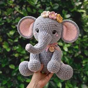 Puede incluir: Un elefante de ganchillo gris hecho a mano con orejas forradas de rosa y una corona de flores. El elefante tiene una cinta rosa alrededor del cuello con una etiqueta de madera que dice "Ellie". El elefante está sentado y tiene una trompa.