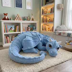 Pode incluir: Um grande dragão de crochê azul claro com chifres brancos e olhos castanhos. O dragão está em um tapete branco em um quarto infantil, cercado por livros e outros brinquedos.