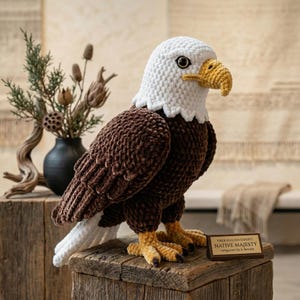 Può includere: Una scultura di aquila all'uncinetto con testa bianca, corpo marrone e becco e artigli gialli. L'aquila è appoggiata su un blocco di legno, con un cartello che recita "Native Majesty". Un vaso nero con piante secche è sullo sfondo.