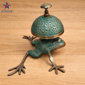 Puede incluir: Una campana decorativa antigua con forma de rana, con una cúpula detallada de color verdín y un pequeño mango redondo. La rana tiene las patas y los pies extendidos, y la campana está montada sobre su espalda. La campana es de metal.