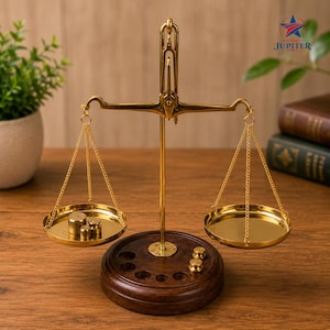 Puede incluir: Una balanza de metal dorado con dos platos circulares suspendidos por cadenas. La balanza está montada sobre una base circular de madera marrón oscuro con ranuras integradas para pesas. Tres pequeñas pesas de metal descansan en el plato izquierdo.